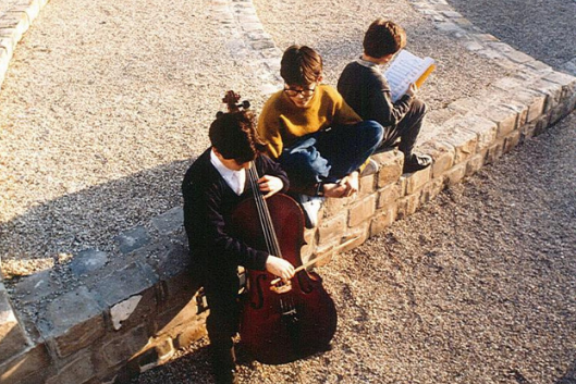 Three youth sitting together; one playing the cello, one watching, and one reading.